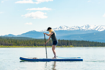 Woman with blonde hair paddle boarding in northern Canada with snow capped mountains in the background in summer time.  - obrazy, fototapety, plakaty