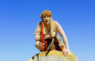 Fototapeta premium A young woman is dressed as Neanderthal warrior. Her body and face is covered with mud and dirt. She is seen posing in a stone quarry. 
