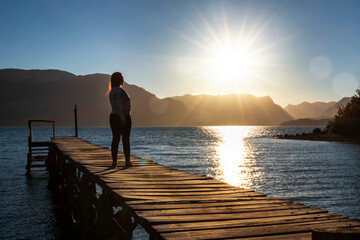Mujer viajera mirando el sol caer desde un muelle de un lago patagónico