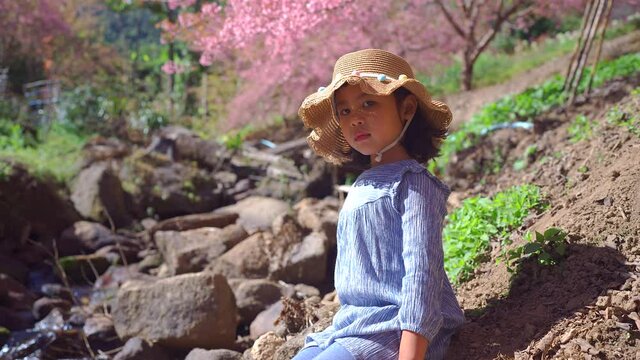 Cute small girl sitting at stream against blossom prunus serrulate flowers.
