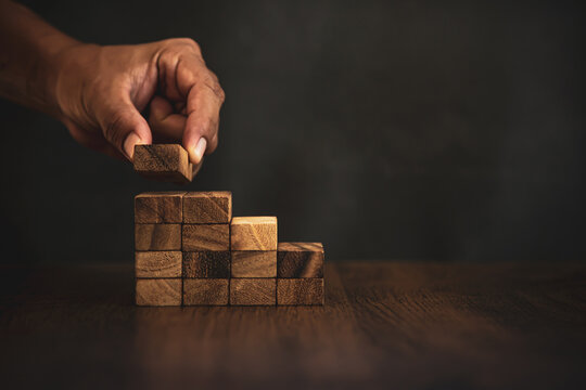 Close-up hand is placing wood block tower stacked in stair step with caution to prevent collapse or crash concepts of financial risk management and strategic planning.