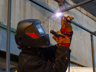 A man in a mask welds the construction. Industrial worker at a welding plant. A strong man - a welder, a welding mask, welds a metal product with arc welding, blue sparks fly to the sides.