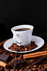 White cup of strong, invigorating black coffee on white saucer on black background with natural roasted coffee beans, pieces of dark chocolate and cinnamon sticks. Tilt horizon, vertical frame