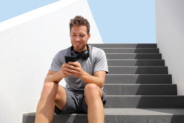 Fototapeta premium Happy man using phone outside relaxing at home or fitness gym listening to audiobook with wireless headphones. Young people lifestyle.