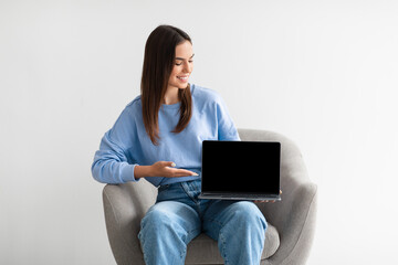 Smiling woman showing laptop with empty screen, sitting in armchair on white studio background, mockup