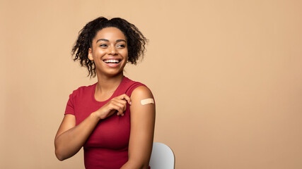 Cheerful vaccinated black woman showing arm with bandage after coronavirus vaccine injection on beige background - obrazy, fototapety, plakaty