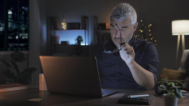 overwhelmed stressed mature man working late at night at the computer from home,caucasian middle aged male at the desk works at the laptop feels tired takes off eyeglasses rubs his eye