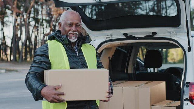 Mature african american smiling man holding cardboard box stands near open trunk of automobile happy elderly male courier outdoors in work clothes accepts goods handyman loader loads boxes delivery