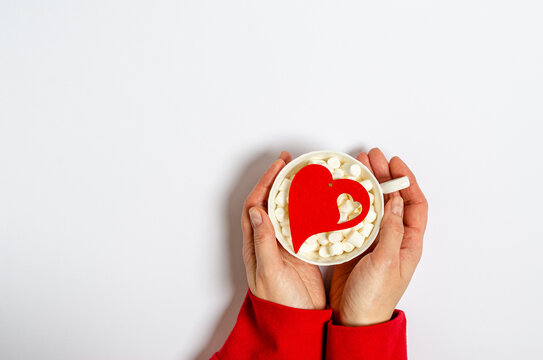 Cup with marshmallows and a big red heart in female hands on a white background with place for text