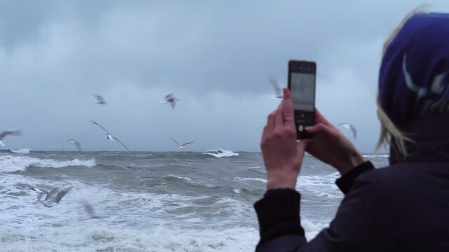 A woman takes pictures of a storm at sea and a lot of birds on her smartphone.