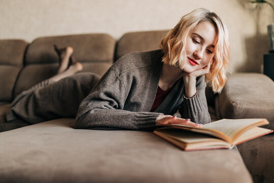 Thoughtful Student Girl Reading Book. Young Woman Studying At Home, Sitting On Beige Couch