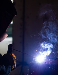A welder in a workshop with a welding machine, welds metal structures. A worker in a factory uses a welding mask, tools and metalworking equipment.