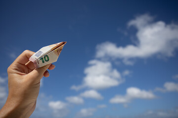 Hand holding the plane made of 10 euros bill on a blue sky with clouds background.