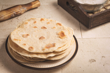 Indian bread (roti) on brown and beige plate, flour in a glass plate in brown wooden tray and wooden brown rolling pin are on beige stone table.
