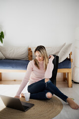 A woman sitting on the floor on a carpet, searches the internet on her laptop.