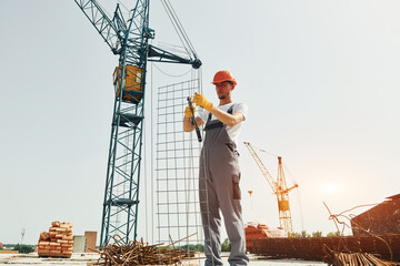 Crane is at background. Young construction worker in uniform is busy at the unfinished building