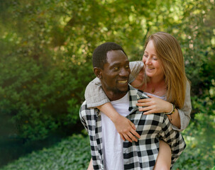 Smiling african man carrying his blonde blue-eyed girlfriend on his back in a green space