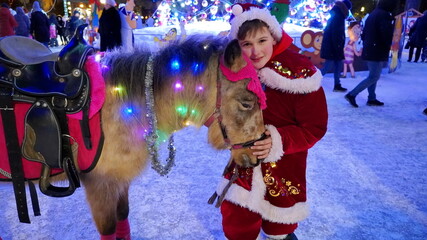 a teenager in a red carnival costume with a pony horse stands near a Christmas tree in a city park...