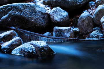 Blue hour, a photo of a mountain stream in the Carpathians in Poland. A huge mass of stones covered with snow and ice. The flowing water turns white and blue. Stone-based branch with ice crystals