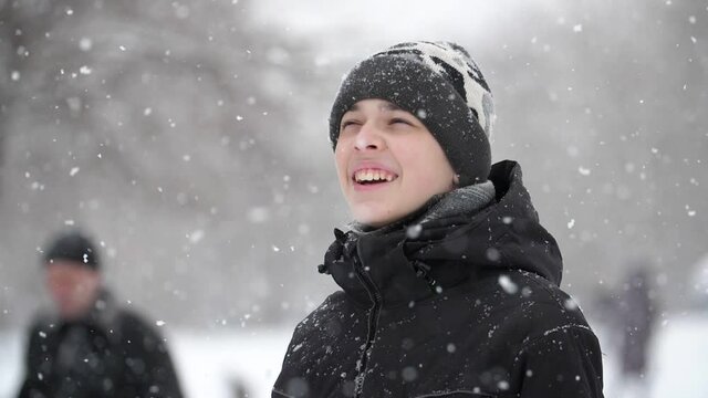 A 13-year-old European boy, dressed in a black jacket, stands outside in a heavy snowfall, he looks at the snow and smiles. Close-up, slow motion, shallow depth of field