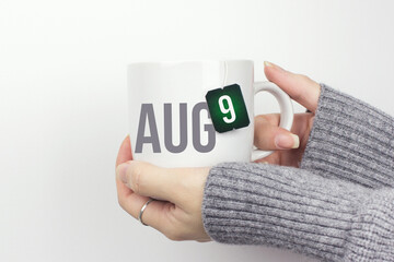 August 9th. Day 9 of month, Calendar date. Closeup of female hands in grey sweater holding cup of tea with month and calendar date on teabag label. Summer month, day of the year concept.