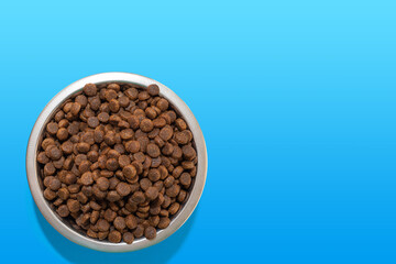 Round metal bowl full of crunchy dry food for dogs or cats on blue background, top view, copy space. Pet care concept. Delicious and healthy food for domestic cats and dogs. Diet food for pets.