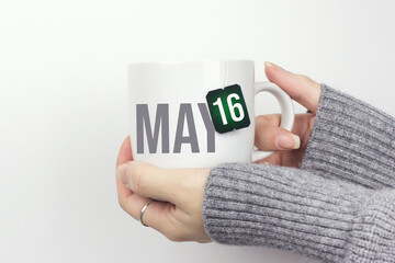 May 16th. Day 16 of month, Calendar date. Closeup of female hands in grey sweater holding cup of tea with month and calendar date on teabag label. Spring month, day of the year concept.