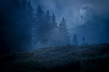 A mystical misty night in the mountains with a full moon and a silhouette of an old wooden cross and dark birds flying in the sky.