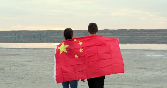 Young man and woman in jeans walk lifting bright Chinese flag above heads and joining hands on calm river bank on windy day backside view