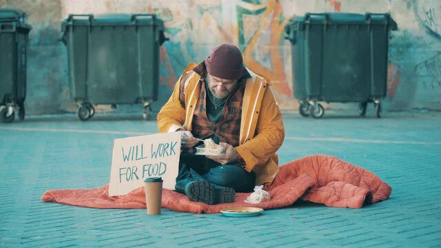 A beggar is eating on the ground near waste bins