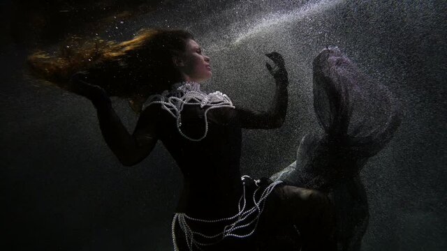 elegant woman in black dress with white pearl is swimming underwater, romantic slow motion shot
