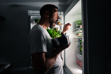 Young hungry man eating food at night and looking in open fridge. Man taking midnight snack from...
