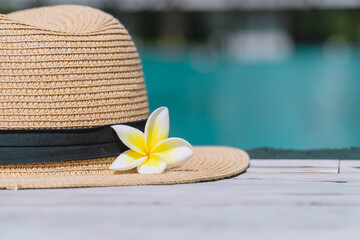 straw hat with plumeria flower on wooden floor with blurred blue water background. summer and holiday concept