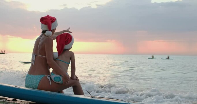 Young brunette mother and preschooler girl in colorful swimsuits and red Santa hats sit on SUP board enjoying sunset and celebrating Christmas