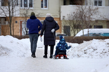 Fototapeta na wymiar Family leisure in winter, mother rides a child on sledge. Couple walking with kid in residential district