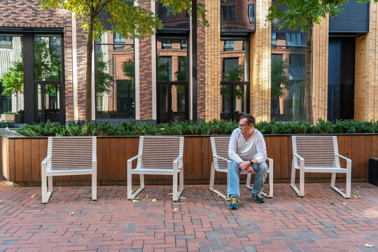 Millennial man in trousers and white T-shirt with long sleeves looking away while sitting on one of four chairs in courtyard of modern brick residential complex with landscaping on city street