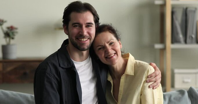 Portrait of grown up son hugs his older happy attractive mother sit on sofa at home smile look pose at camera. Bonding and affection, family ties, connection between two gen relatives people concept