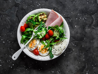 Delicious balanced breakfast, snack bowl - ham, boiled egg, grilled zucchini, green salad in one bowl on a dark background, top view