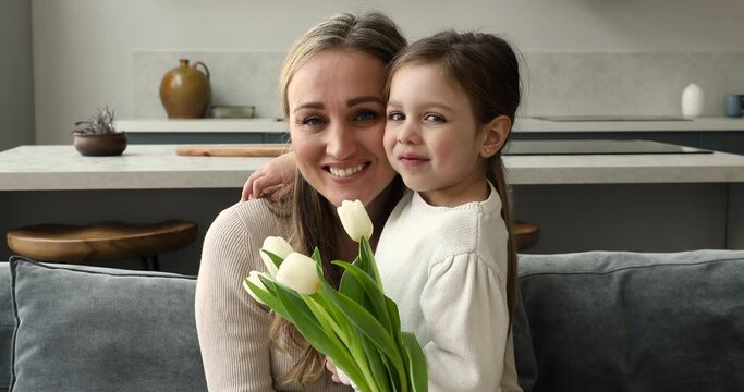Beautiful woman holding white bouquet fresh tulips hugs little 4s daughter look at camera. Loving family celebrate Happy Mother Day or birthday feeling love and affection, life events congrats concept