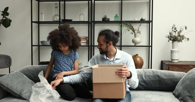 African girl, her father sit on couch unpack parcel box with fragile kitchenware. Concept of satisfied clients of e-commerce retail e-services, trusted transport company, express delivery services