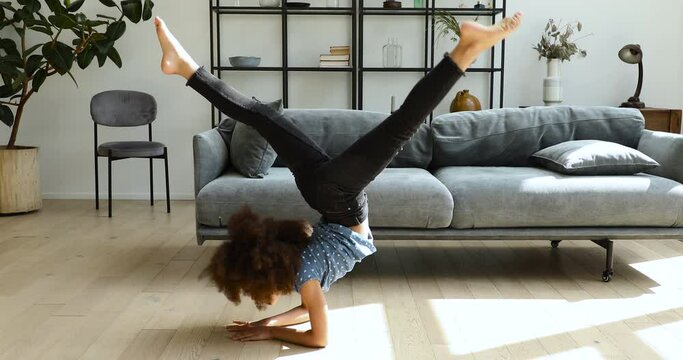 Cute little 10s African curly haired girl practising yoga, perform acrobatic poses throws her legs over head while stand on elbows in living room. Gymnastic work out at home, active lifestyle concept