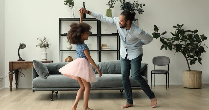 Cute little African girl wear fluffy festive pink skirt swirling while holds loving daddy hand, dance together in living room. Family leisure, love and bond, Father Day, life event celebration concept