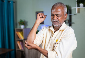 indian senior old man exercising by strenching hand due to elbow joint pain at home - concept of...