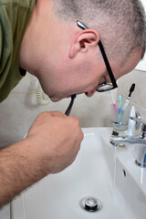 A white man with glasses is brushing his teeth in a bathtub above the sink. The tap is open and water is flowing.