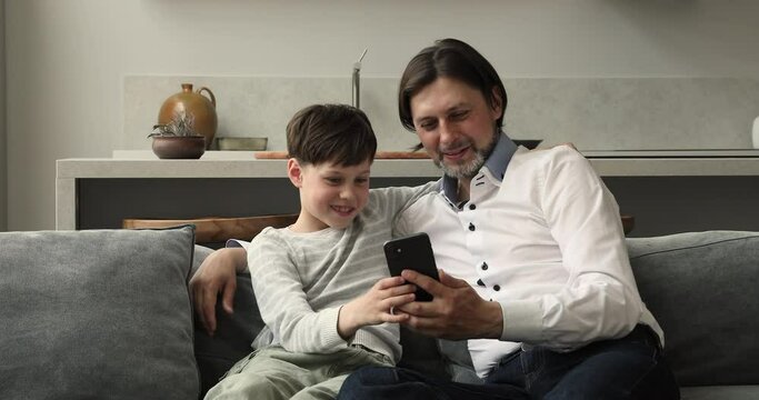 Dad and 8s son use together smartphone sit on sofa at home, play new cool online game, enjoy mobile application on cellphone. Young gen use modern tech, fun and kid development, device overuse concept
