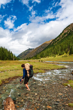 Young man with big backpack barefoot crossing river. Shoeless male backapcker in river. Karakol valley, Issyk-kul region, Ala-kul lake Terskey Alatau mountain range, Kyrgyzstan, Central Asia.