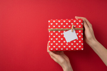 First person top view photo of st valentine's day decor girl's hands demonstrating giftbox in red wrapping paper with heart pattern twine and pinned note on isolated red background with copyspace