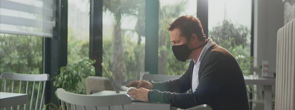 Caucasian male working on laptop in empty cafe. Wearing protective face mask to avoid spreading Covid-19. Entrepreneur working outside office.