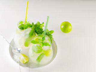 Mojito cocktail with lime and mint on white background. Two glasses of mojito with straws. Summer refreshing cocktail and bar accessories on a white plate. Copy space