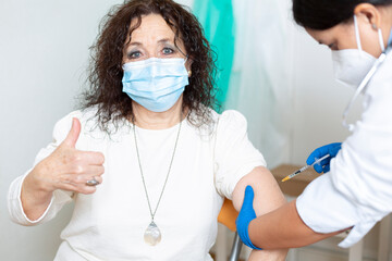 Older caucasian woman with her thumb up while being vaccinated by a female medical worker. Coronavirus vaccination concept.